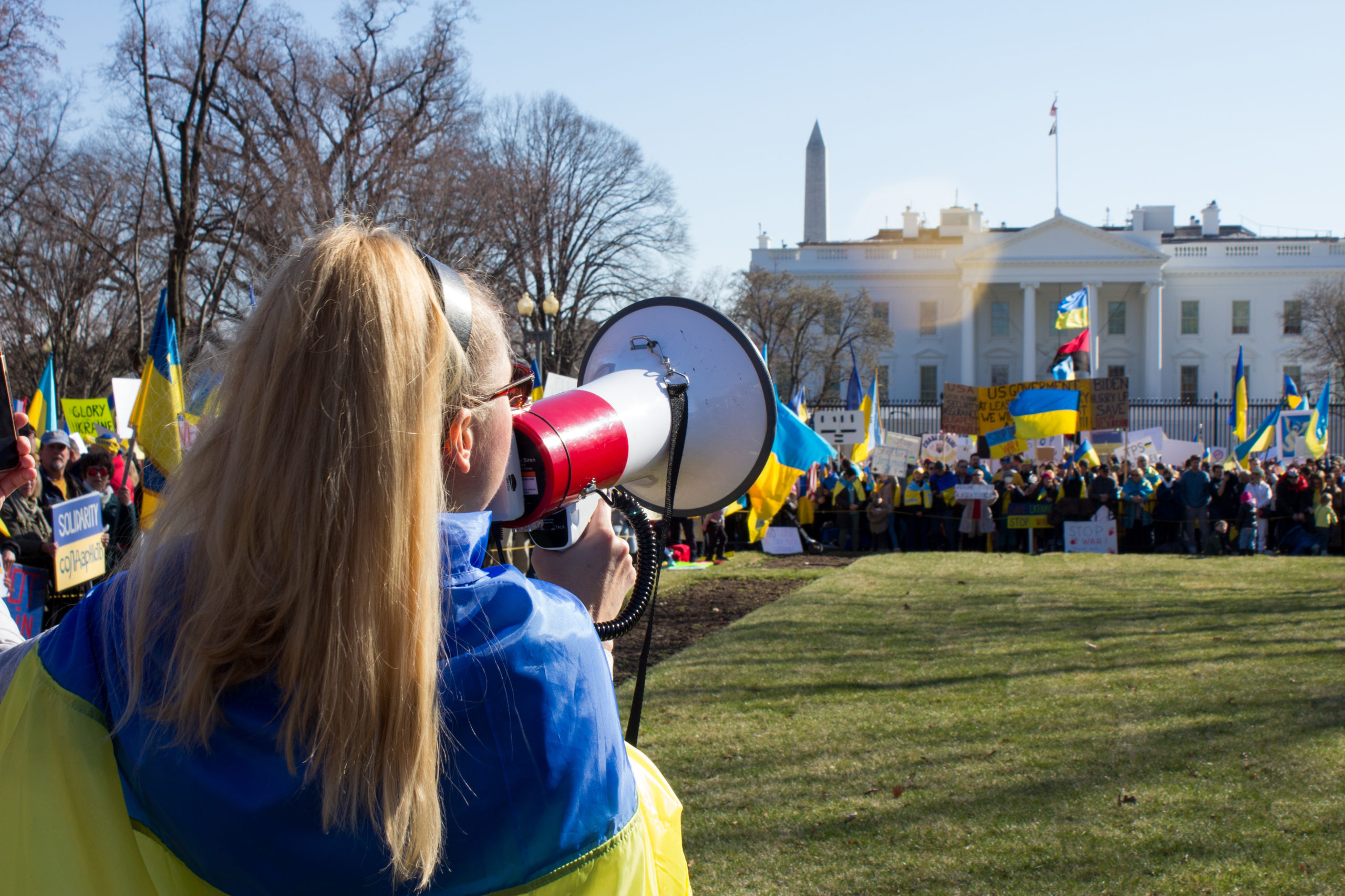 A woman with a megaphone and a Ukrainian flag speaks in front of a crowd outside the White House